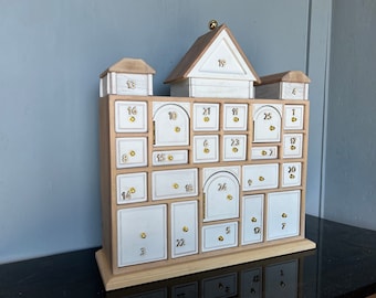 May include: A wooden advent calendar with 25 numbered drawers, each with a small door. The calendar is shaped like a house with a roof and a chimney. The numbers are in white and the doors are white with gold accents.