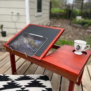 Könnte beinhalten: Ein Laptopständer aus Holz mit roter Oberfläche und schwarzem Lederbezug. Der Ständer verfügt über einen integrierten Tablethalter und einen kleinen Beistelltisch. Auf dem Beistelltisch steht eine weiße Tasse mit schwarzem Design.