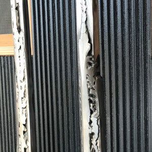 May include: Two tall, silver metal sculptures with a textured, rough surface. The sculptures are standing upright against a black, ribbed background.