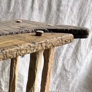 Puede incluir: Dos bancos rústicos de madera con patas talladas y superficies texturizadas. Uno es de color claro, mientras que el otro es más oscuro, mostrando la veta natural y los nudos de la madera. Los bancos parecen hechos a mano.