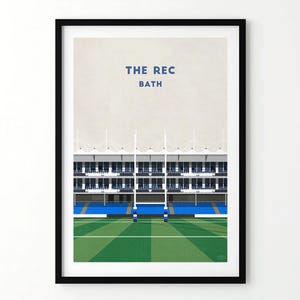 Puede incluir: Una impresión enmarcada que presenta una ilustración minimalista de un estadio deportivo. La imagen muestra un campo verde, asientos azules y el texto "THE REC BATH" en azul. La arquitectura del estadio se representa con líneas limpias y un fondo neutro.
