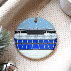 May include: A round ceramic ornament featuring a stadium design. The ornament has a light blue sky with white snowflakes at the top and a stadium with blue and white seating. A gold string is attached for hanging.