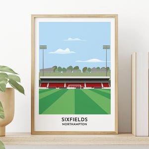 Peut inclure: Illustration numérique du stade de Sixfields à Northampton, en Angleterre. Le stade est représenté dans un style minimaliste avec un terrain vert, des sièges rouges et un ciel bleu avec des nuages blancs. Le texte "SIXFIELDS NORTHAMPTON" est imprimé sous le stade.
