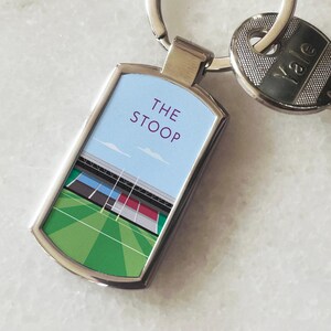 Peut inclure: Un porte-clés en métal argenté avec une étiquette rectangulaire représentant une illustration colorée d'un stade de rugby. L'étiquette porte le texte "THE STOOP" imprimé dessus.