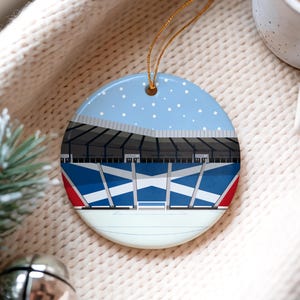 Può includere: Ornamento natalizio rotondo in ceramica con un design a stadio con dettagli blu e bianchi, che ricorda una bandiera. La parte superiore presenta un cielo blu con fiocchi di neve bianchi. Un filo d'oro è attaccato per appenderlo. Una decorazione festiva.