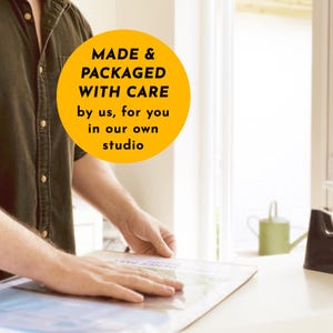 May include: A person's hands carefully packaging a print. A bright yellow circle with the text "MADE & PACKAGED WITH CARE by us, for you in our own studio" is in the upper left corner. The print is on a white surface.