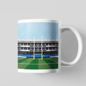 May include: White ceramic mug with a stadium design. The image shows a sports field with green grass, blue details, and a grandstand under a light blue sky. A great gift for sports enthusiasts.