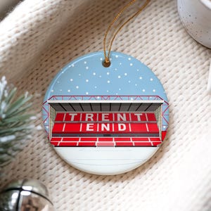 Può includere: Ornamento natalizio rotondo in ceramica con un design a stadio e la scritta "TRENT END" in rosso. L'ornamento ha uno sfondo azzurro chiaro con dettagli di fiocchi di neve bianchi e un cordino dorato per appenderlo.