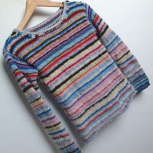 May include: A multi-coloured striped long-sleeved sweater, displayed on a wooden hanger. The sweater features a round neckline and ribbed cuffs. The stripes include shades of blue, red, yellow, and grey. The garment is made of knitted fabric.