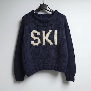 Könnte beinhalten: Ein dunkelblauer, langärmeliger Strickpullover mit einem dicken gerippten Kragen und Bündchen. Das Wort SKI ist in großen, cremeweißen Blockbuchstaben in der Mitte der Brust eingestrickt. Das Kleidungsstück hängt an einem schwarzen Kleiderbügel.