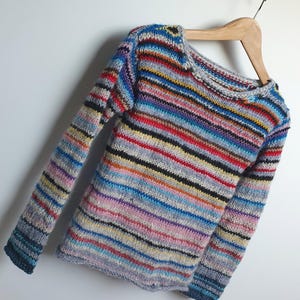 May include: A crew neck sweater with long sleeves, displaying a vibrant array of horizontal stripes in colours such as blue, red, and yellow. The knitted garment is hung on a wooden hanger against a white backdrop. The knitwear appears to be handmade.