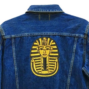 Könnte beinhalten: Eine blaue Jeansjacke mit einem goldenen gestickten Pharaonenkopf-Patch auf der Rückseite. Die Jacke hat einen Knopfverschluss und zwei Taschen auf der Vorderseite. Der Text "24 Planets Studio" befindet sich in der unteren rechten Ecke der Jacke.