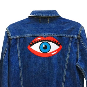 May include: A blue denim jacket with a red and blue eye patch on the back. The patch has a white eyelash and a black pupil. The text "24 Planets Studio" is on the bottom right corner of the jacket.