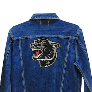 Puede incluir: Una chaqueta vaquera azul con un parche de pantera negra en la espalda. El parche tiene un contorno plateado y ojos amarillos. La chaqueta tiene cierre de botones y dos bolsillos en la parte delantera.