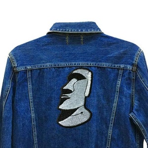 May include: A blue denim jacket with a black and white embroidered patch of a face on the back. The text "24 Planets Studio" is on the bottom right of the jacket.