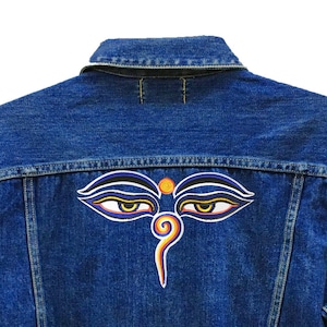 Puede incluir: Una chaqueta vaquera azul con un diseño de ojo bordado de colores en la espalda. El texto "24 Planets Studio" está en la esquina inferior derecha de la chaqueta.