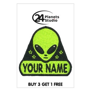 Pode incluir: Rosto de alienígena verde com olhos pretos e um triângulo preto com estrelas brancas ao redor do alienígena. O alienígena está em uma faixa preta com o texto "YOUR NAME" em verde.
