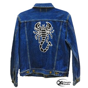 Pode incluir: Uma jaqueta de ganga azul com um remendo de escorpião preto nas costas. O remendo é um design simples com contornos brancos.