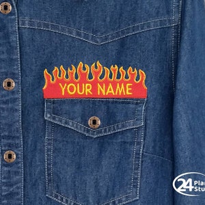 May include: A dark blue denim shirt featuring a red and yellow flame patch with the text "YOUR NAME". The shirt has a button-down design, two chest pockets, and brass-coloured buttons. The patch is embroidered.