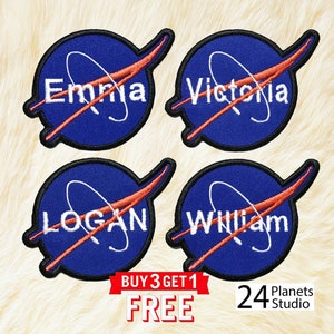 May include: Four blue and orange embroidered iron-on patches with the names "Emma", "Victoria", "Logan", and "William" inside a white circle with a red swoosh. The patches are on a light brown fuzzy surface. The text "BUY 3 GET 1 FREE" is in a red banner below the patches. The text "24 Planets Studio" is in black text below the banner.