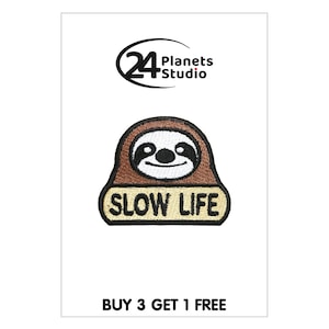 May include: Brown and white embroidered patch featuring a sloth face and the text "Slow Life" in black lettering on a beige background. The patch is outlined in black.