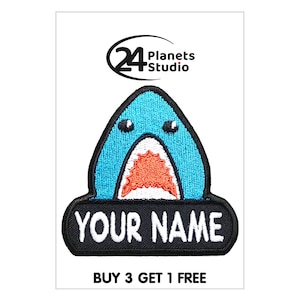 May include: Embroidered patch with a cartoon shark design. The shark is blue with white teeth and an orange mouth. The patch includes the text "YOUR NAME" on a black banner and the "24 Planets Studio" logo.