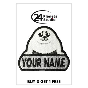 May include: White and black embroidered iron-on patch featuring a cartoon seal holding a banner with the text "YOUR NAME". The patch is from 24 Planets Studio.
