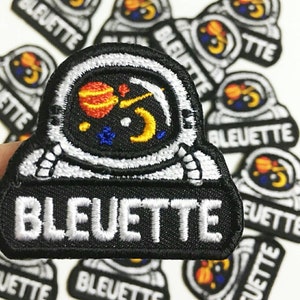 Puede incluir: Un conjunto de 10 parches termoadhesivos que representan a un astronauta de dibujos animados con planetas en su casco. Los parches son negros con bordados blancos y de colores. El texto "BLEUETTE" está bordado en blanco en la parte inferior de cada parche.