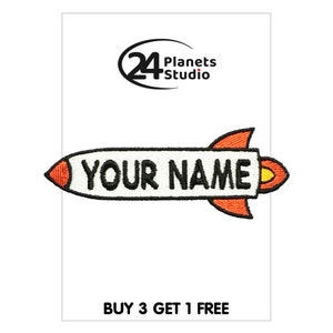 May include: White iron-on patch shaped like a rocket with a black outline and orange flames. The patch says "YOUR NAME" in black text. The patch is for clothing and is sold by 24 Planets Studio.