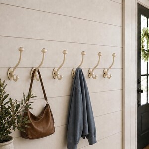 May include: A row of six cream-colored metal wall hooks mounted on a white shiplap wall. A brown leather shoulder bag hangs from the second hook, and a blue sweatshirt hangs from the fourth hook. The hooks feature a curved design with ball ends.