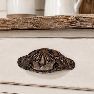 May include: A semi-circular, dark bronze-toned metal drawer pull with an embossed shell pattern, mounted on a rectangular, distressed off-white wooden drawer front with a natural wood-toned top edge and visible screw heads.