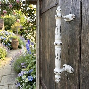 May include: A white metal door handle with a distressed finish featuring visible dark patches of wear. The handle has a vertical, elongated shape with decorative rounded accents at the top, middle, and bottom, mounted onto a dark wooden door.