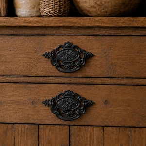 May include: A wooden cabinet featuring two drawers with ornate black metal pulls. The wood has a visible grain and a warm brown finish. The drawers are stacked vertically, each centered with a detailed, oval-shaped black metal handle.