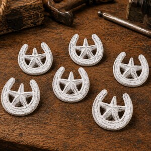 May include: Six white, distressed-finish metal ornaments, each shaped like a horseshoe with a five-pointed star centered within the arch. The items feature a textured, weathered appearance with visible white paint over a darker base material.