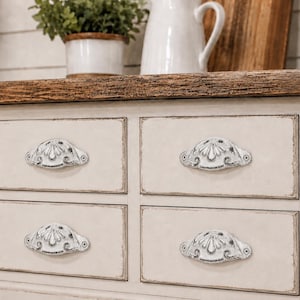 May include: A small wooden chest of drawers featuring a distressed off-white finish and a natural dark wood top. The unit has four rectangular drawers, each equipped with a decorative, ornate, white metal cup pull handle with dark accents.