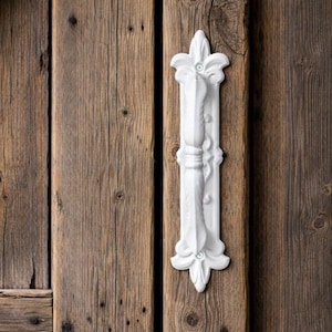 Peut inclure: Une poignée de porte blanche et ornée, avec un motif floral, est fixée sur une porte en bois vieilli. La poignée comporte une barre verticale avec des éléments décoratifs et une poignée centrale. Le bois a un aspect naturel et vieilli.