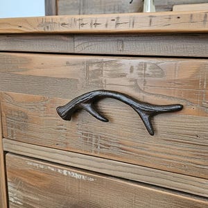 May include: Close-up of a rustic wooden dresser with a dark, antler-shaped drawer pull. The dresser has a natural wood grain finish with visible knots and imperfections, giving it a distressed look. The drawer pull is made of metal.