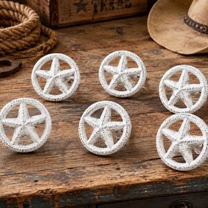 Könnte beinhalten: Ein Set aus sechs runden Schrankknöpfen, die jeweils ein fünfzackiges Sternmotiv innerhalb eines Rings aufweisen. Die Knöpfe haben eine abgenutzte, strukturierte weiße Oberfläche, die ihnen ein verwittertes Aussehen verleiht.