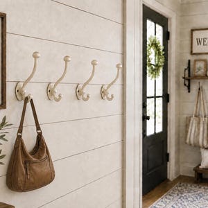 May include: Four off-white metal wall hooks with a curved, double-prong design mounted on a horizontal shiplap wall. Each hook features a circular base plate with visible screw holes, providing a functional storage solution for hanging items.