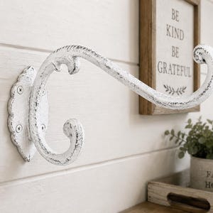 May include: A wall-mounted coat hook with a distressed white finish and a curved, ornate design. The metal hook features a scalloped mounting plate secured by two visible screws, showcasing a weathered texture throughout its entire structure.