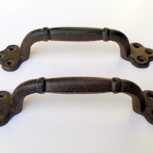 May include: Two black cast iron door pulls with a simple design. Each pull has a rounded handle and a decorative clover-shaped end.