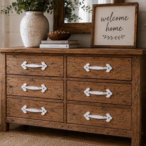 Puede incluir: Una cómoda de madera con seis cajones, con un acabado marrón desgastado y tiradores de metal blanco ornamentados. La parte superior cuenta con un jarrón de cerámica blanca, un cuenco de madera, dos libros apilados y un letrero enmarcado con el texto welcome home.