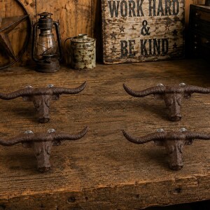 Könnte beinhalten: Ein Set aus vier braunen Wandhaken aus Gusseisen, die jeweils wie ein Longhorn-Stierkopf mit markanten, geschwungenen Hörnern geformt sind. Die Haken haben eine texturierte, verwitterte Oberfläche und sind in einem zwei-mal-zwei Raster angeordnet.