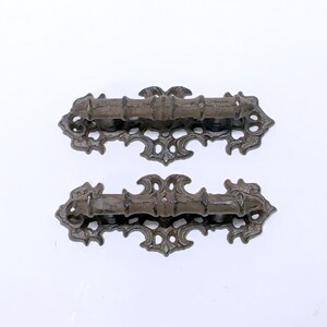 Puede incluir: Dos tiradores de caj&oacute;n de metal marr&oacute;n oscuro adornados con intrincados dise&ntilde;os de volutas. Los tiradores tienen una forma rectangular con un asa ligeramente curvada.