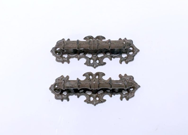 Puede incluir: Dos tiradores de caj&oacute;n de metal marr&oacute;n oscuro adornados con intrincados dise&ntilde;os de volutas. Los tiradores tienen una forma rectangular con un asa ligeramente curvada.