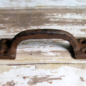 May include: A rustic brown cast iron door handle with a square shape and two holes on each side. The handle is distressed and has a worn finish.