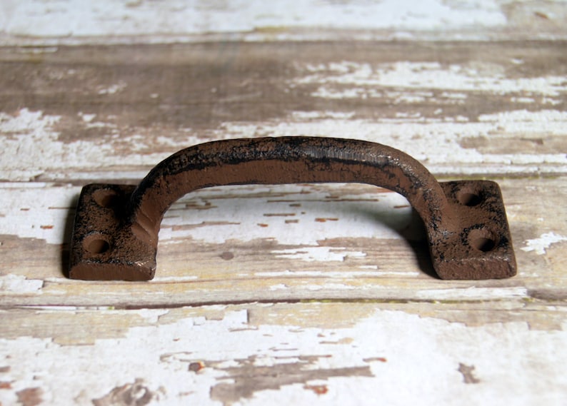May include: A rustic brown cast iron door handle with a square shape and two holes on each side. The handle is distressed and has a worn finish.