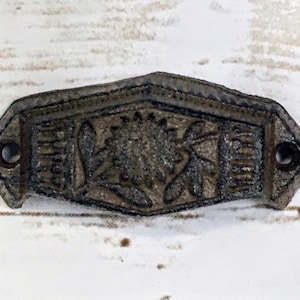 May include: A cast iron drawer pull with a floral design. The pull is a dark gray color and has a slightly distressed finish.
