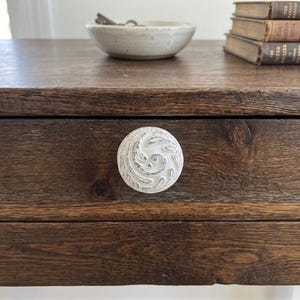 Puede incluir: Primer plano de un cajón de madera marrón oscuro con un pomo de cerámica blanco con remolinos. El cajón forma parte de una pieza de madera más grande, con un cuenco pequeño y libros visibles encima. El pomo es el punto focal.