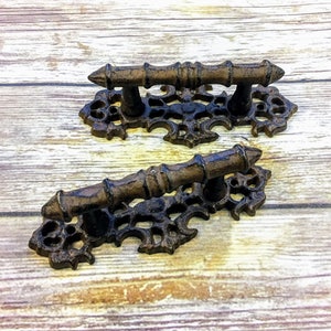 May include: Two ornate cast iron drawer pulls with a decorative scroll design and a brown finish. The pulls have a rectangular base and a short, straight handle.